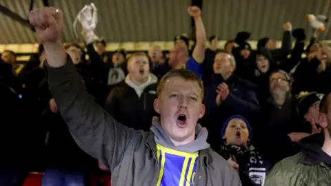 Getty Images Weston-super-Mare FC fans cheer their team on from the stands at Blundell Park during the FA Cup tie with Grimsby Town. In the front is a young man with a grey hoodie over which he is wearing a yellow and blue striped Weston top. Some fans are holding small versions of the FA Cup