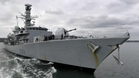 Royal Navy FOST Members of the crew of HMS St Albans standing on its deck. The warship, seen here sailing, is painted light grey and has the hull number F83. It is equipped with various antennae, radar systems, and a large gun turret on the deck. The background shows a cloudy sky and a coastline in the distance. 