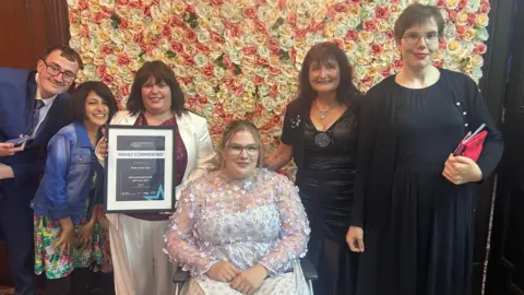City of Wolverhampton Council A group of five women and one man are in front of a flower wall. The woman in the middle is sat in a wheelchair and the woman to the left of her holds up a certificate.