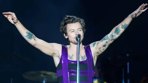 Harry Styles in a purple sequinned and black vest and tattoos on both arms - with two birds on his chest - he holds his hands up in the air and sings into the microphone