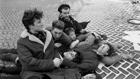 Beezer A group of six teenagers lying on the ground with various big coats and shirts and one wearing sunglasses. They are in an artistic huddle on the stone-bricked pavement.