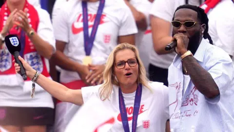 Getty Images Sarina Wiegman puts an limb astir Burna Boy arsenic he performs on-stage successful beforehand of a statement of England players astatine nan team's triumph celebrations.