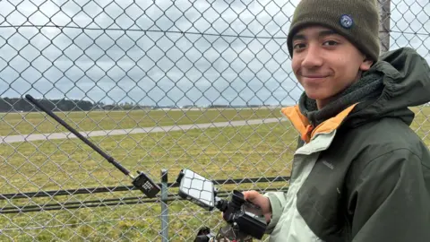 Matt Precey/BBC Teenager in a beanie hat holding a camera. He is smiling. Behind him is a fence and an airfield. Attached to the fence is a radio scanner.