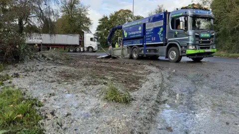 The scene at Bolham Roundabout in Tiverton, with a large tanker positioned on the roundabout and a lorry behind it. The road and land around the roundabout appear to be soaked in fluid.