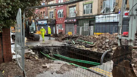 A hole in the road with green cables over it - surrounded by metal fencing. Workers in yellow outfits and white hats stand nearby