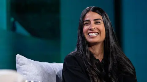 Getty Images A woman with long dark hair sits on a sofa on-stage at a business event. She looks relaxed as she smiles at the host off-camera.