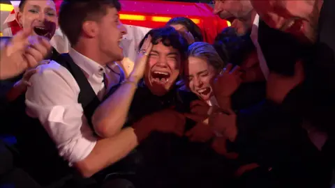 JJ clutches his head as he is surrounded by people