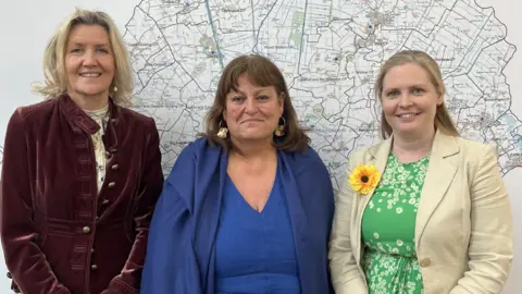 Three women smile at the camera. Behind them is an ordnance survey map of Huntingdonshire. 