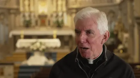 Fr Phil Sumner talks to camera. He has white hair and is wearing a clerical collar with a V-necked jumper over the top. He sits with a church altar blurred in the background