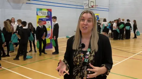 Lucy Moore a teacher at Trinity Secondary school stood in a school hall wearing a lanyard around her neck and talking to the camera as school pupils gather around behind her talking while holding their new green backpacks