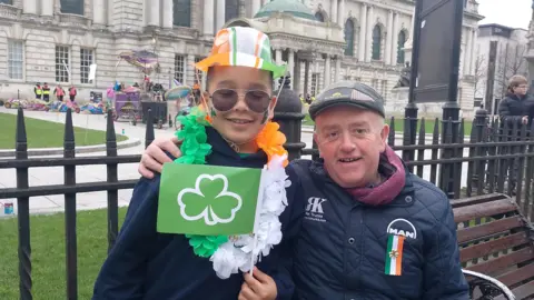 Seorang anak dan seorang pria yang lebih tua digambarkan di bangku di luar balai kota Belfast. Anak itu mengenakan topi plastik hijau, putih dan oranye, karangan bunga putih dan oranye hijau dan mengibarkan bendera shamrock hijau. Pria itu mengenakan topi datar, jaket angkatan laut dan mengenakan lencana bendera Irlandia yang disematkan dengan shamrock emas. 