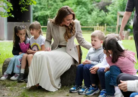 PA Media The princess sits on a log with two children on her left and three to her right. She has her hand the back of one boy who has short fair hair and is dressed in a grey top and navy blue jeans and trainers