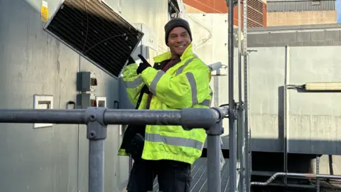 Bristol Old Vic Mike Elliot wearing black cargo trousers, a yellow hi-vis coat, brown beanie hat and black gloves. He is standing on a metal scaffolding platform on the side of the Bristol Old Vic theatre, holding up what looks like a vent panel in one hand and pointing at it with the other, while smiling at the camera. 