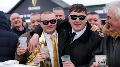 PA Media Two young men pose for a photo among a crowd of people outside a Guinness tent. One wears a gold jacket at tie with black glasses with the words "RIP ME" and "My stag" written on the lenses. The second wears a black jacket and tie and black glasses with the words "Jack's Stag" on the lenses. 