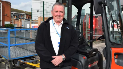 Somerset NHS Foundation Trust Steve Watts is standing in front of a porter trolley. He is wearing a black fleece and a blue NHS lanyard. He is smiling at the camera.