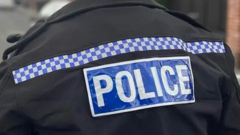 The back of a black police jacket with 'Police' written in white on a blue background. A strip of chequered blue and white squares runs across the top of the jacket.