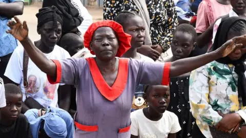 A crowd of Catholic women and children kneeling on the ground outdoors during a Good Friday procession in Juba. the woman standing in the front centre, has her hands up in the air. Other people have lowered heads and gazes, while some appear to be looking ahead