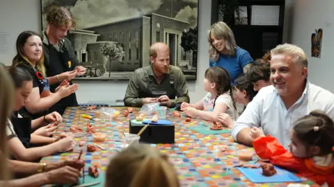 PA Media Prince Harry at a table at the Australian National Veterans Arts Museum (Anvam) in Southbank, Melbourne, Victoria - he is wearing a dark green shirt and is talking to a child, with a woman standing next to them and smiling - there are other people gathered around the table, which has a colourful table cloth
