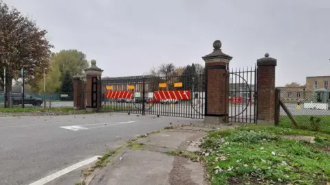 The gates to RAF Scampton. Four brick pillars and black gates with red and orange signs on. There is a road leading up to the gates which is outlined by path and grass. 