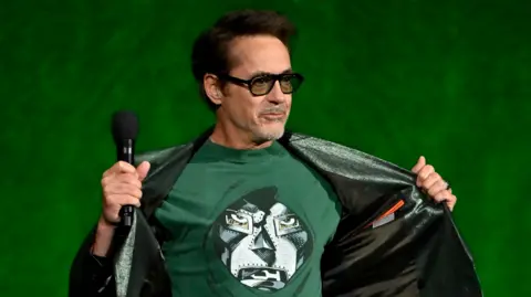Robert Downey Jr on stage at CinemaCon, opening his jacket to reveal a T-shirt showing the masked face of his character Doctor Doom