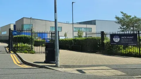 An image of a school, which is a big white building with "Jack Hunt School" written on it in silver, and black gates in front of a car park.