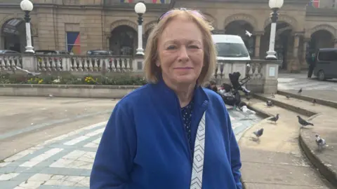 Anna Jameson/BBC June Forest, 70, who has shoulder length light brown hair and is wearing a blue fleece, smiles at the camera on a stone square.