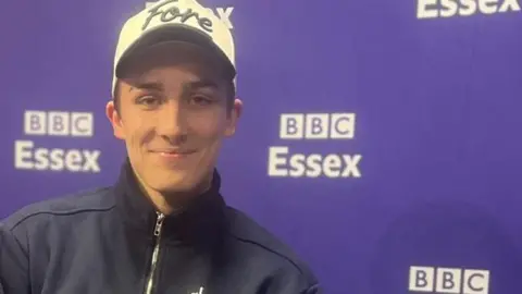 James Patient/BBC Isaac Elles is wearing a navy jumper and a white cap. He is smiling while sitting in a radio studio.