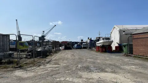BBC Griffiths Yard - part of it is behind barriers, there is a gravel roadway in the middle and buildings on the right with a boat and some cars