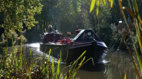 The Coventry canal in Fradley, Lichfield. A man steers a barge through the water on a sunny day. The embankment is lined with trees and greenery and the sun bounces off the water.