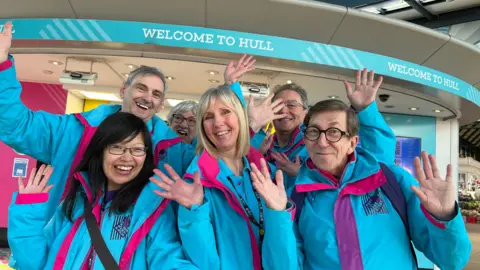 Three women and three men smile and hold up their arms in celebratory fashion in front of "welcome to Hull" signs at a railway station. They are wearing bright blue and pink jackets. 
