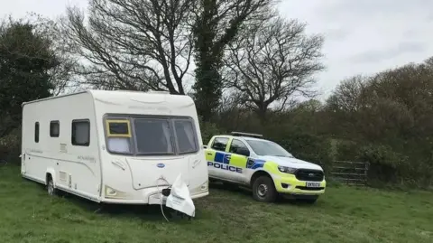 A police 4x4 parked next to a caravan in a field. It is a grey day, with the trees behind bare of leaves. The caravan has dark windows and has nothing outside it.
