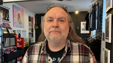 Ellen Knight/BBC Mark Fulton is looking into the camera - he is wearing a black T-shirt with a black, white and red checked shirt over the top. He has long, light brown hair and a short grey beard. He is photographed inside his shop, which has a black back wall, with the two walls framing him painted white. On the walls, and on shelves, are rows and rows of vinyl records for sale. 