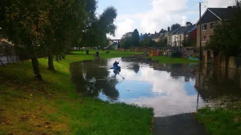 Pip Jones A kayaker paddles through floodwater at a park in Plymouth. The large volume of water is covering a chunk of the park. Green trees line the side of the green grass. Several homes are overlooking the park.