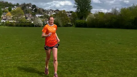 A woman wearing an orange t-shirt and running shorts is running across a grass recreational ground. 