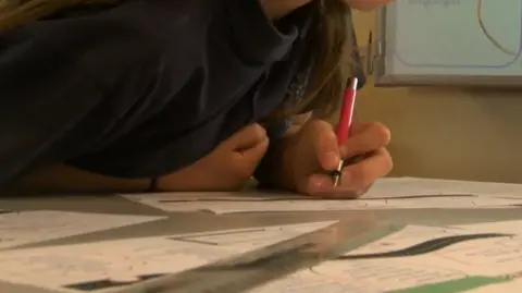 BBC A child leans over a desk to write on a piece of paper. Other sheets are on the desk as well. There is a screen with some information on in the background.