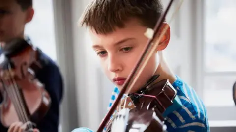 A boy playing the violin, with dark hair and wearing a blue and white striped t-shirt