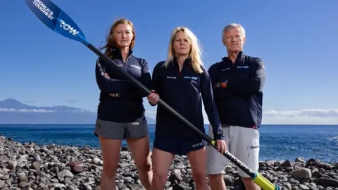 Two women and a man stand on a pebble beach holding an oar and looking at the camera. They wear identical tops and shorts. The woman on the left has long brown hair, the one in the middle long blond hair and the man on the right has short grey hair.