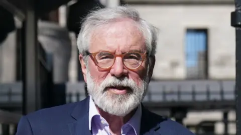 Gerry Adams with short grey hair and facial hair. He is wearing a navy suit with a light purple shirt. He has round, orange glasses on. Behind him is black gates.