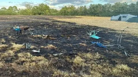 Scorched grassland with burnt remnants of camping chairs and a portable barbecue. A wheelbarrow stands next to the barbecue. A green tent is nearby in the field.tent 