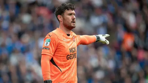 Getty Images Jed Steer wearing an orange Peterborough kit with sponsor 'Mick George' written on the front. He is pointing to the left with a white goalie glove on his hand. There is a blurred crowd of fans behind him.