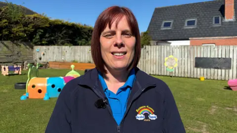 Alison Toole has a bob hiarcut which is red/brown in colour. She is wearing a light blue polo shirt with a dark brown fleece over the top which has the Rainbow Corner logo - a rainbow - on the left breast. She is standing in the outdoor area at the pre-school, behind her various pieces of outdoor play equipment can be seen and further back is a wooden fence.