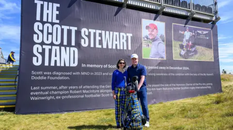 PA Media Scotland's Robert MacIntyre and Robyn Stewart pose in front of a large stand which has a poster informing people that it has been dedicated to Scott Stewart who died of MND in December 2024. It features two photographs of Mr Stewart and three paragraphs of text background about him. The description mentions his love of golf and notes that he died from the condition in December last year. 