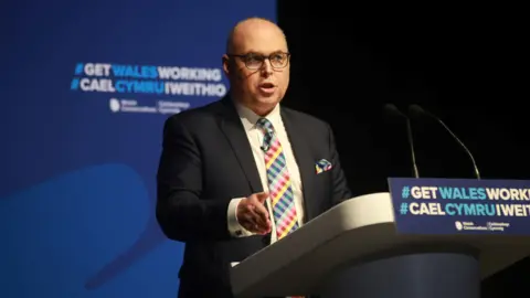 Welsh Conservatives Paul Davies stood giving a speech, pointing with his hand down to a lectern which has the words "Get Wales Working" written in English and Welsh attached to it on a blue background.