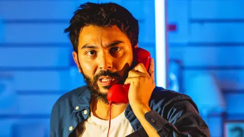 Andrew Billington A cast member looking anxious and holding a red old-style phone receiver to his ear