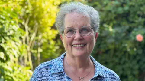 A woman with grey hair and glasses is smiling at the camera. She has a blue top and bushes are blurred in the background.