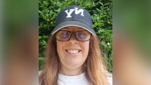 Jo Willis A selfie-style image of Jo Willis. She is smiling at the camera as she stands outside in front of a large green bush. She wears a navy cap, black glasses, a white top and has long brown hair.