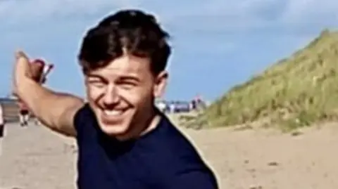 A young man with dark hair and a dark blue t-shirt smiles as he points behind him while standing on a beach.
