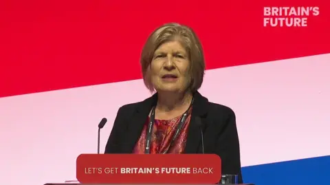 Sally Keeble standing at a podium with a red, white and blue striped background. A message on the podium reads 'Let's get Britain's future back". 