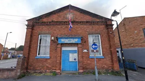 Google A large red brick build with baby blue doors. The building has a flag pole above the large wooden doors with a union flag hanging from it