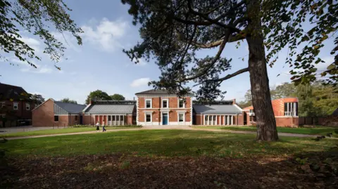 Paul Karalius A red brick building with white windows and grey slanted roof which is surrounded by green space and large trees.